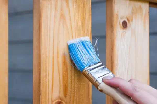 Application d'un produit pour protéger le bois extérieur. Une main tient un pinceau bleu et blanc pour appliquer le traitement sur des poteaux en bois clair.