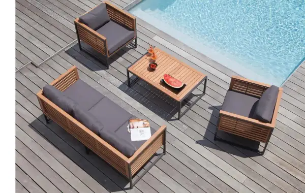 Salon de jardin en bois teck avec coussins gris, près d'une piscine. Table basse avec fruits et boissons.
