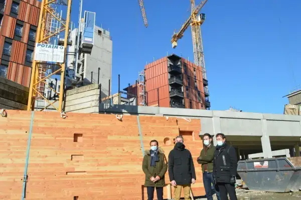 Trois hommes devant un mur en bois, un objet à faire au tour à bois, sur un chantier avec des grues.
