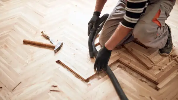 Un artisan utilise un aspirateur pour nettoyer la poussière de bois après le ponçage d'un parquet en chevrons. La pâte à bois blanchie au ponçage est prête pour la finition.