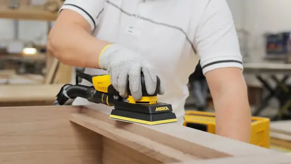 Main gantée utilise une ponceuse Mirka pour poncer une porte en bois, préparant la surface pour une finition parfaite.