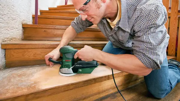 Un homme utilise une ponceuse pour rénover les marches d'un escalier en bois.