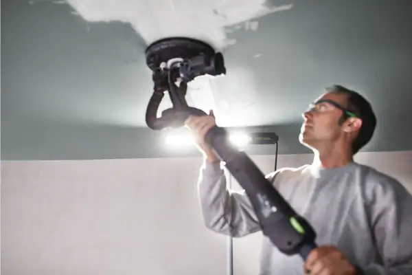 Un homme utilise une ponceuse de plafond Festool, éclairée par une lampe rasante Festool, pour lisser une surface.