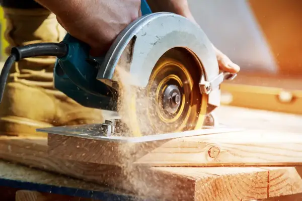 Main d'un artisan guidant une scie circulaire. Le sens de rotation de la lame de scie circulaire projette de la sciure.