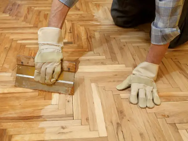 Un homme, ganté, applique de la cire sur un parquet en chevrons avec une raclette.