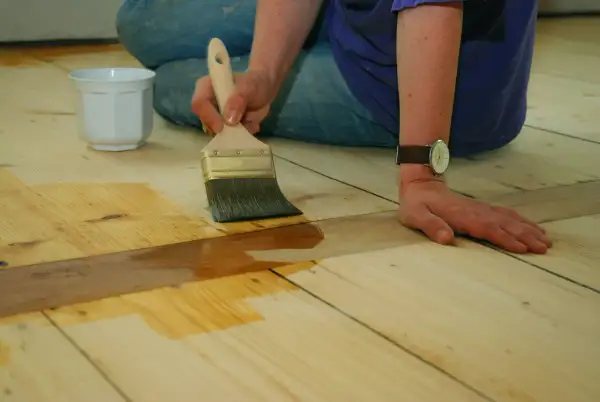 Application d'une couche protectrice sur un parquet huilé ou ciré. Une main tient un pinceau, l'autre est posée sur le bois.