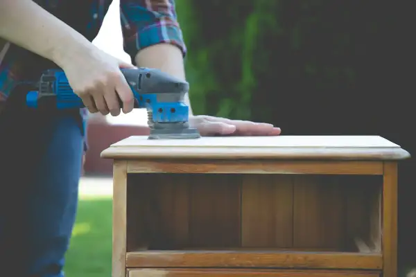 Une personne utilise une ponceuse pour lisser le dessus d'une petite commode en bois, un travail qui demande de poncer endroit difficile.