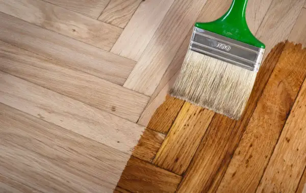 Un pinceau applique une couche de vernis sur un parquet en bois clair, transformant sa teinte. Le processus de nettoyer parquet est en cours.