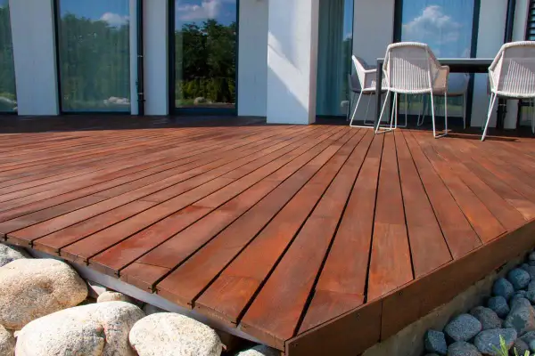 Belle terrasse bois sur plot béton, avec des galets et des gros rochers. Des chaises et une table sont visibles au fond.