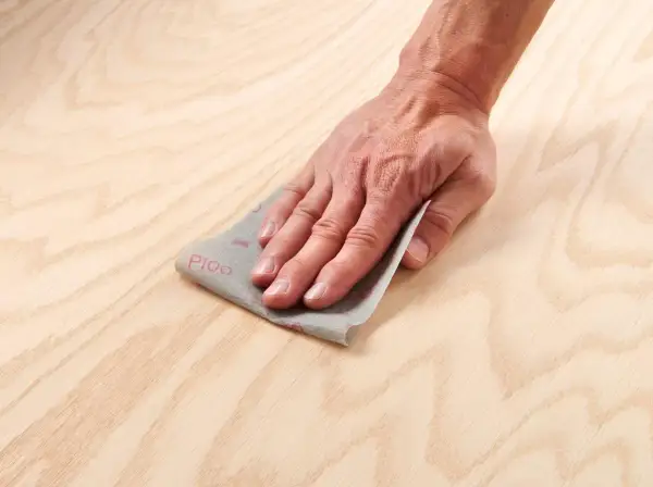 Main d'artisan effectuant un égrenage sur une surface en bois clair avec du papier de verre P100.